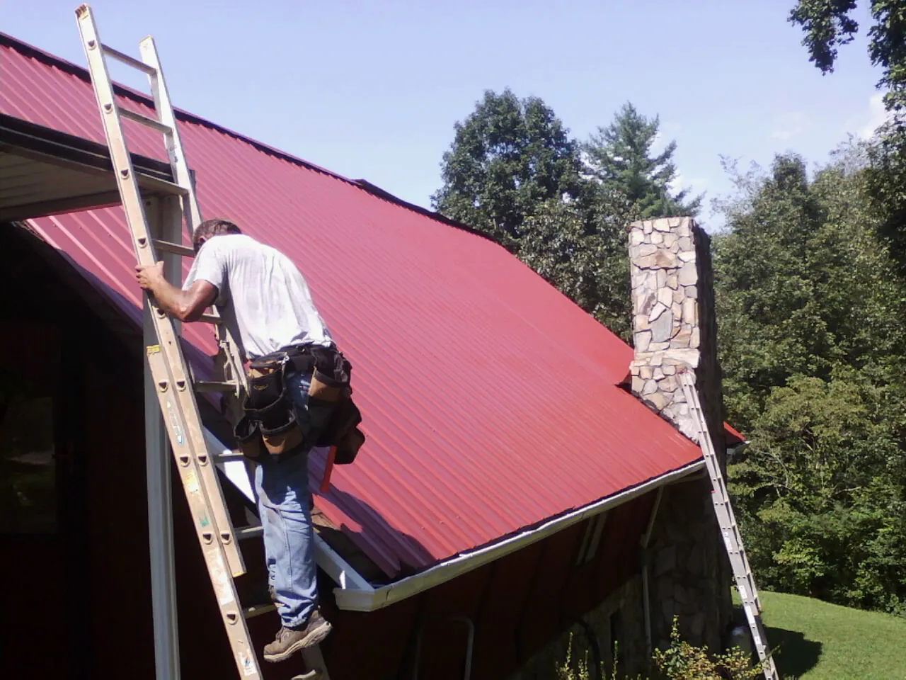 Roofer on a ladder beside a red metal roof and stone chimney — Soffit & Fascia Repair by Roofing Brothers in New Germany